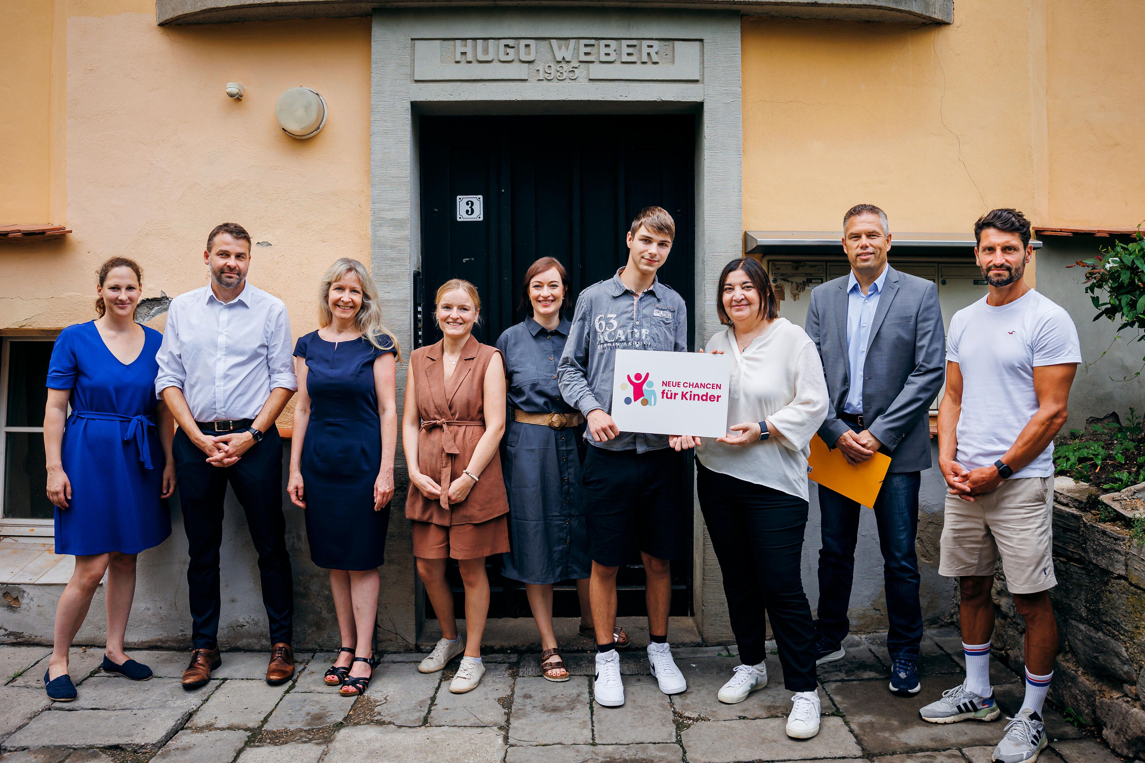 Neun Erwachsene stehen vor einem Haus, zwei davon halten eine Schild mit der Aufschrift: NEUE CHANCEN für Kinder.